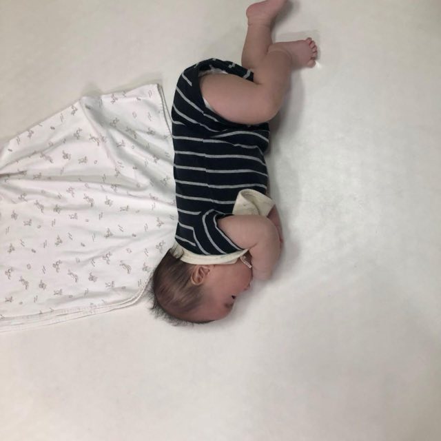 マットに仰向けで寝て脚を上げ、体を横に向けている百日ごろの赤ちゃん