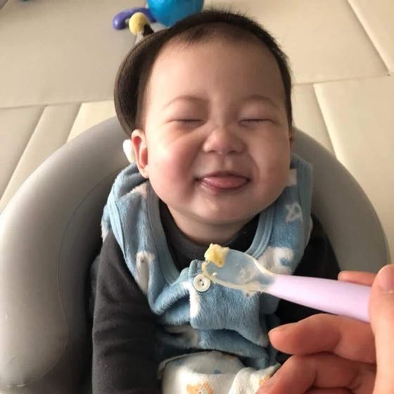 턱받이를 하고 숟가락으로 첫 이유식을 받아먹으며 활짝 웃는 아기