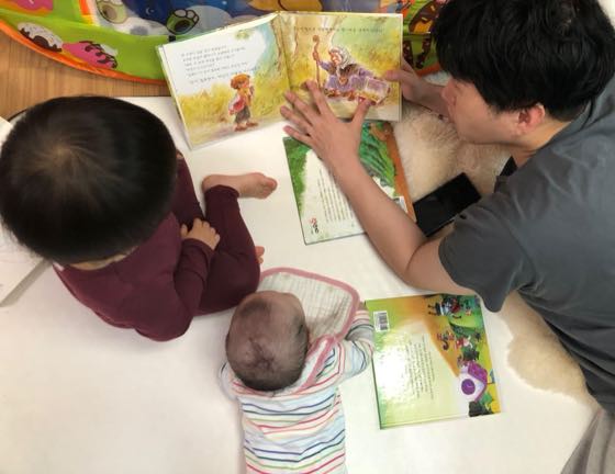 お父さんが絵本を開いて読み聞かせ、赤ちゃんがそばで一緒に絵本を見つめている様子