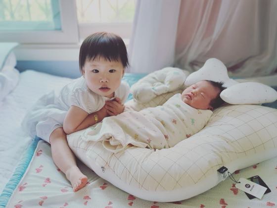 かごのクッションに包まれて眠る生後100日ごろの下の子のそばを、上の子が見守る様子